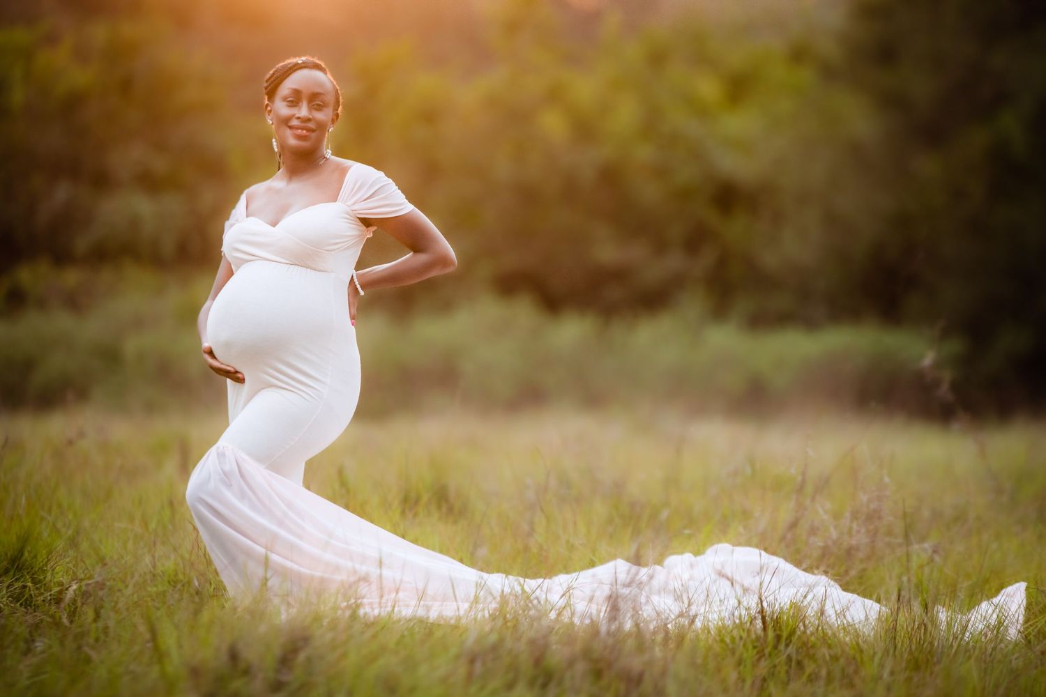 Maternity photos of woman in white dress posing gracefully in sunlit outdoor field.