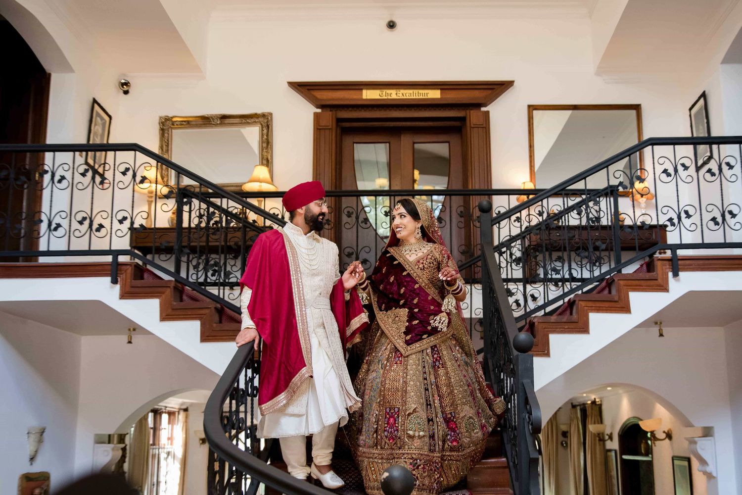 Wedding couple in traditional Indian attire pose on elegant staircase with ornate railings.