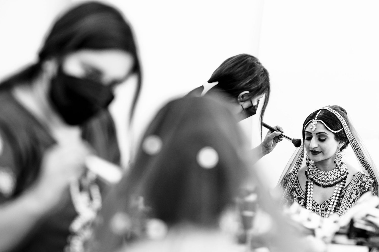 A black and white photo showing preparation of an Indian bride wearing traditional bridal jewelry and attire.