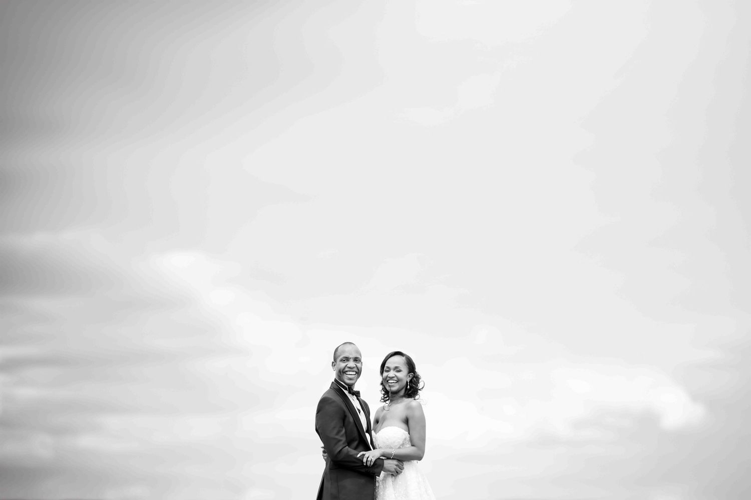 A black and white wedding portrait capturing a joyful moment against a cloudy sky.
