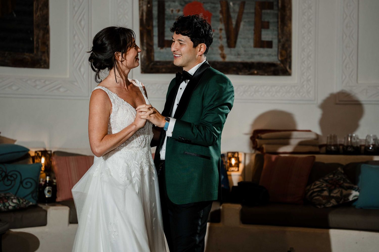 A couple in formal wedding attire dance together in a dimly lit room with elegant decor.