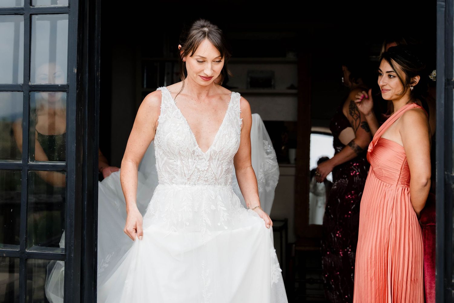 A group of bridesmaids in coral dresses stand with a bride in a white lace wedding gown during a formal event.
