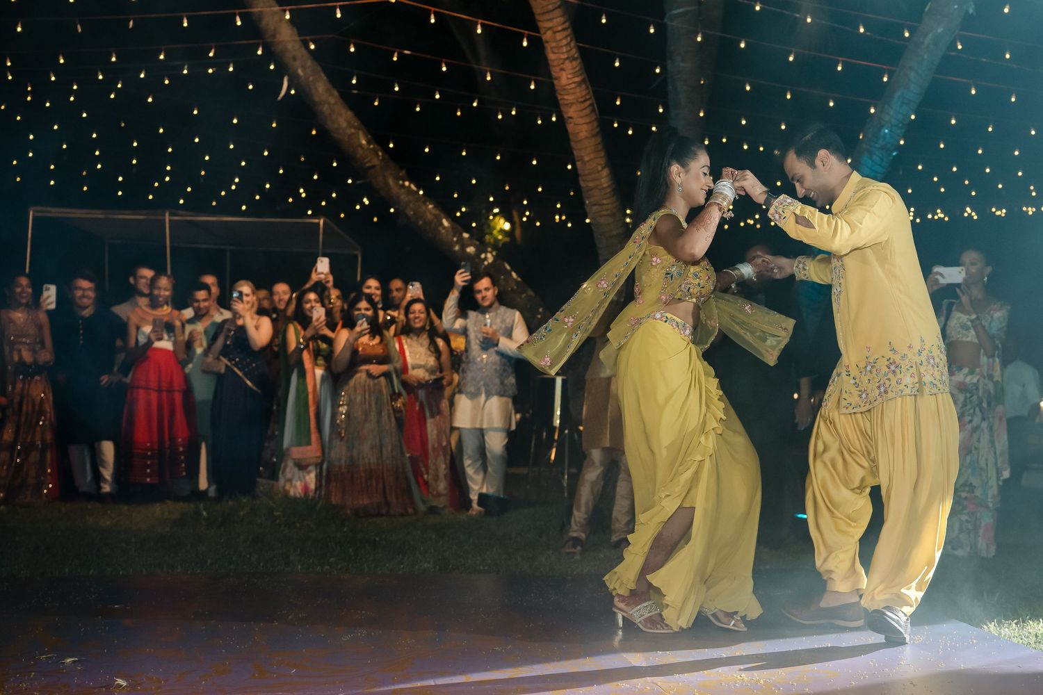 Dancers in yellow outfits perform under fairy lights at an outdoor evening event.