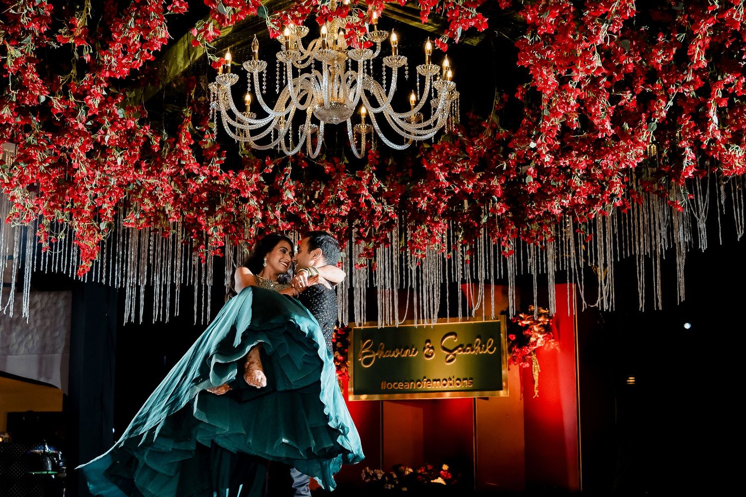 A couple dances beneath a glamorous chandelier and red floral decorations at an elegant wedding reception.