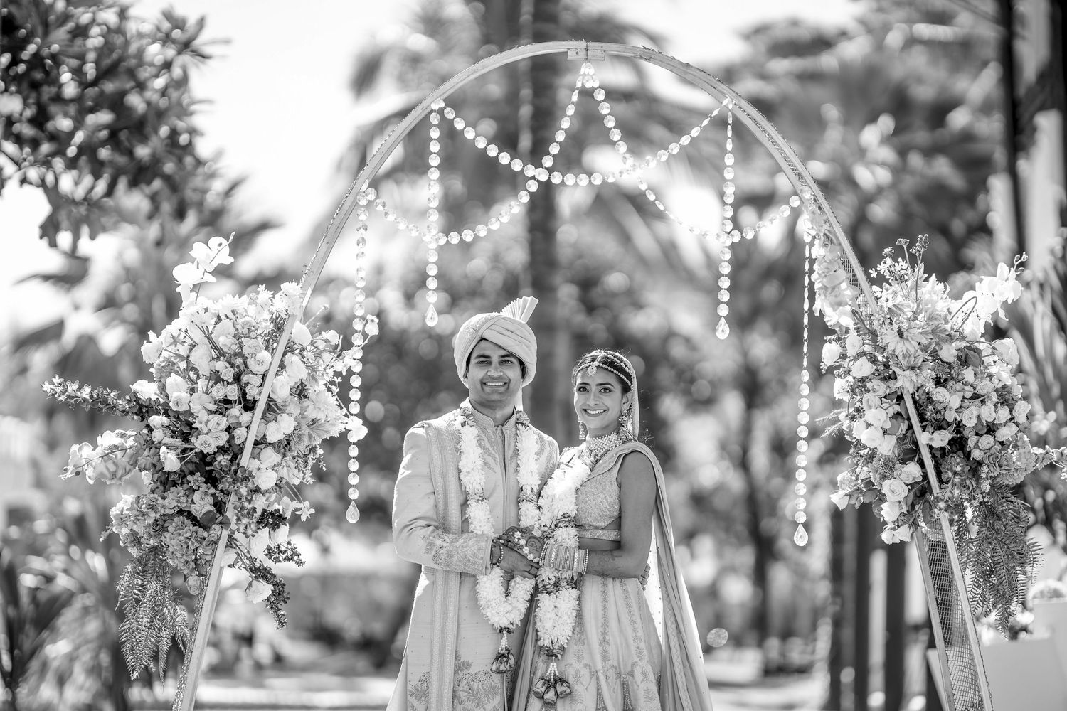 Black and white portrait of couple under decorative wedding arch with string lights.