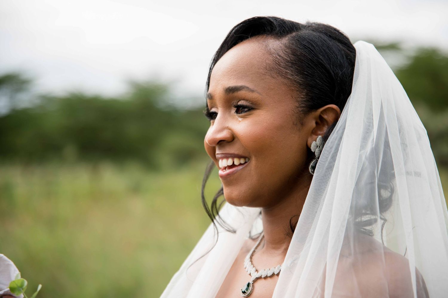 A radiant bride wearing a white gown and veil smiles during an outdoor wedding ceremony.