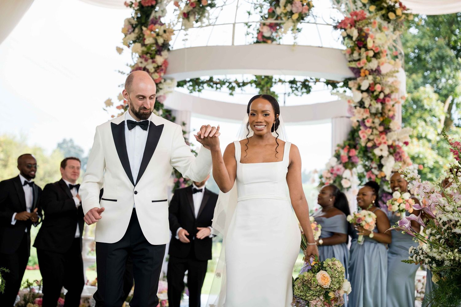 Wedding celebration under a floral arch decorated with pink roses and greenery during an outdoor ceremony.
