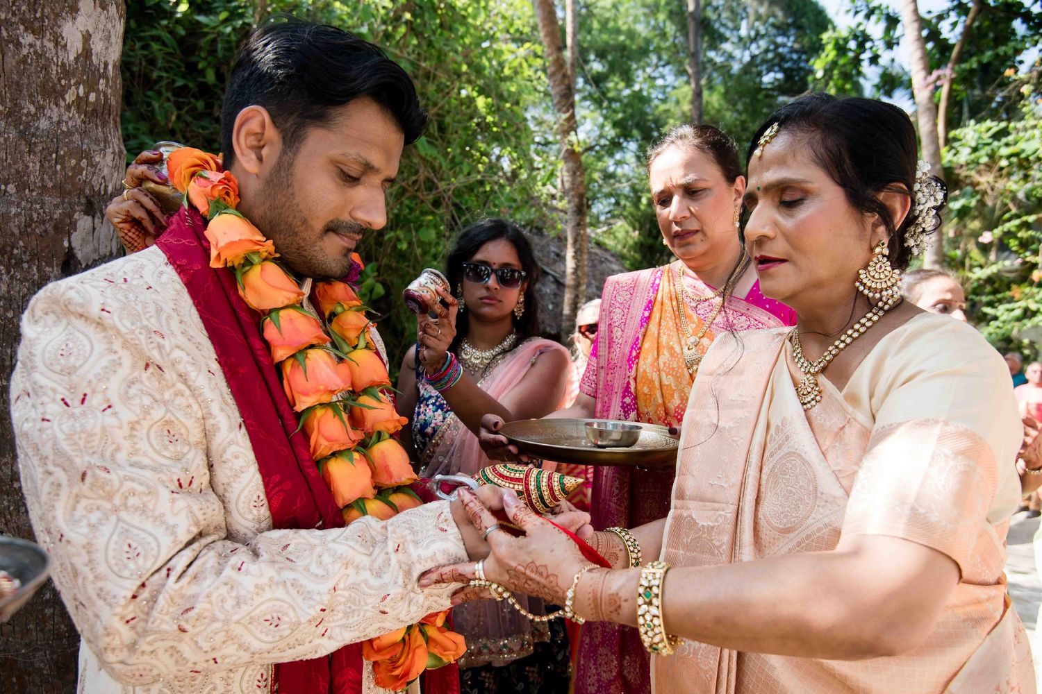 A traditional Indian wedding ceremony takes place outdoors with participants wearing floral garlands and formal attire.