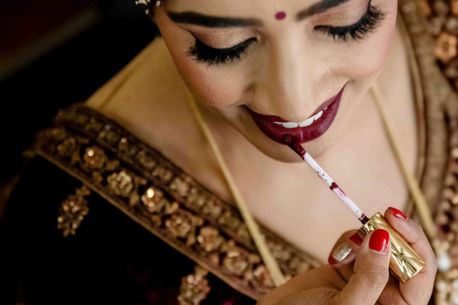Close-up detail of bride applying deep red lipstick showing elaborate Indian bridal jewelry and makeup.