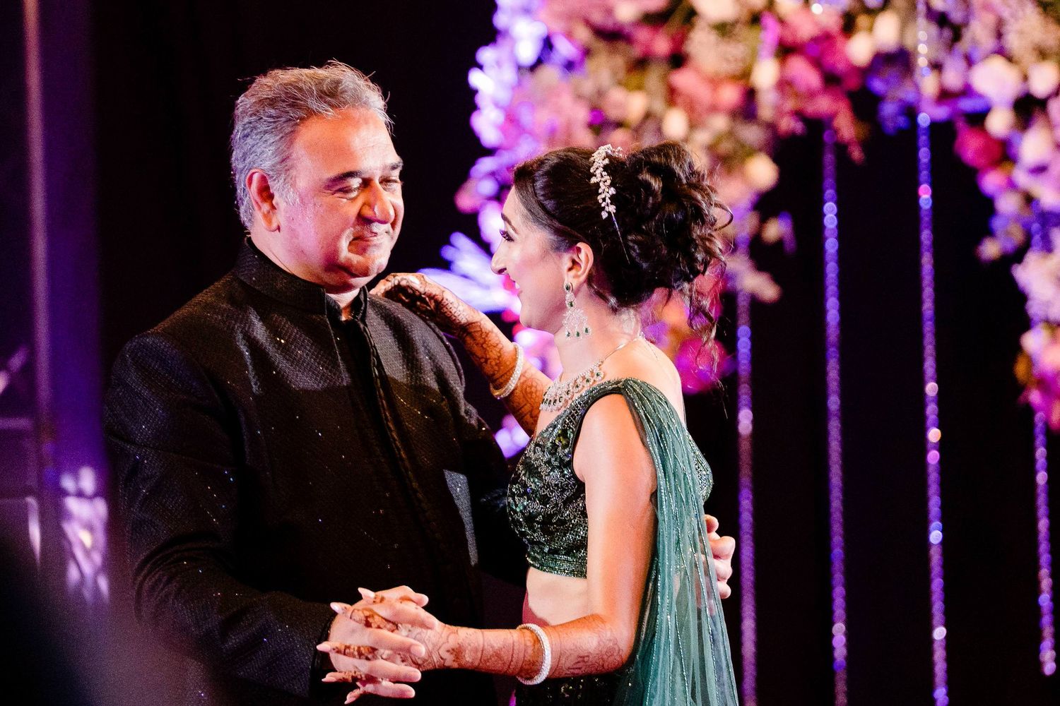 A couple dances together at a formal event with purple lighting and hanging decorations in the background.