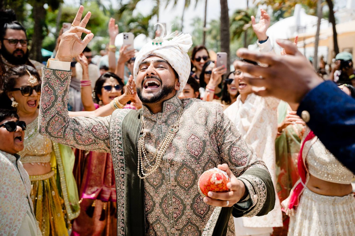 A joyful celebration at an outdoor Indian wedding with guests wearing traditional attire and dancing in the sunlight.