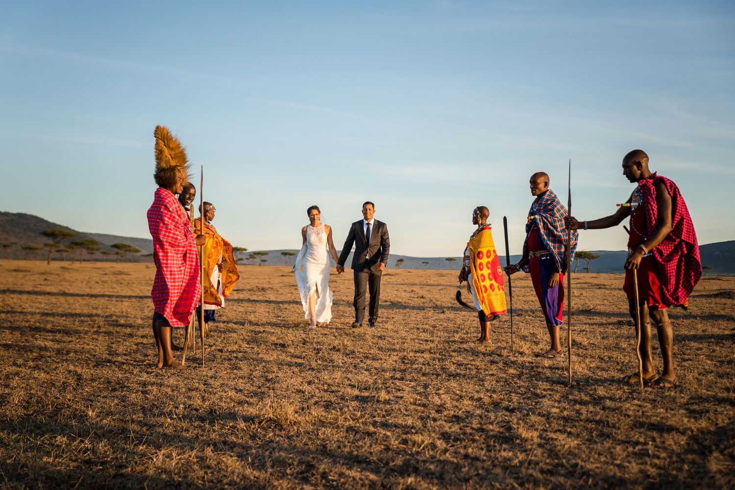People in colorful traditional African attire walking across a golden savanna landscape at sunset.