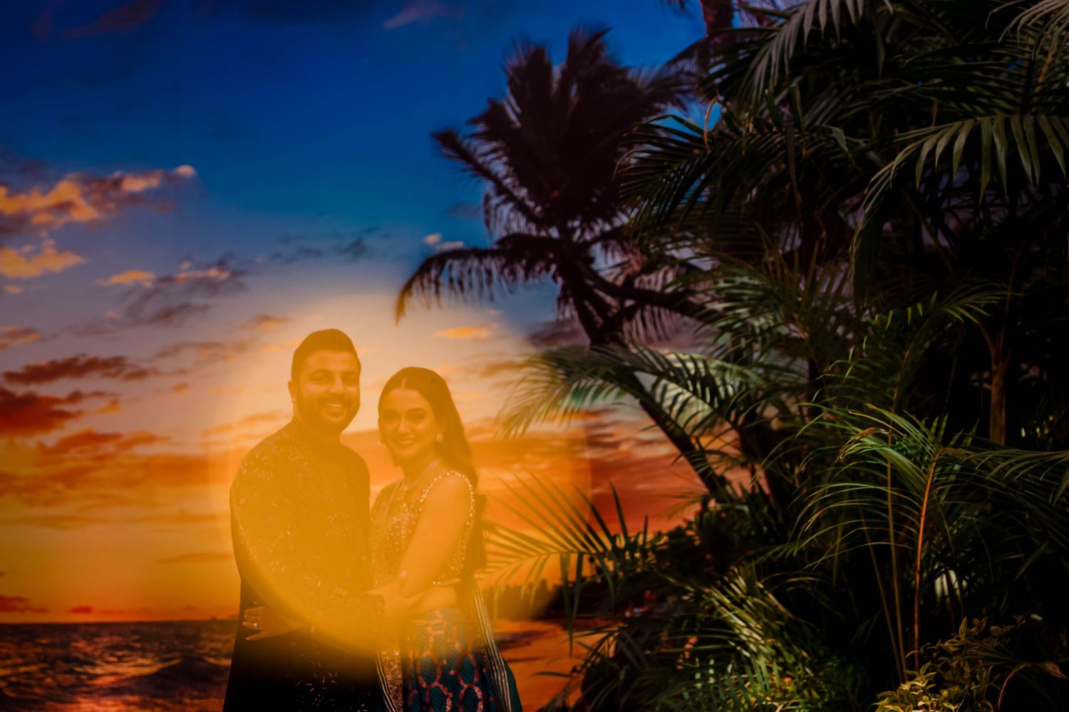 A silhouetted couple embraces against a vibrant sunset with palm trees.