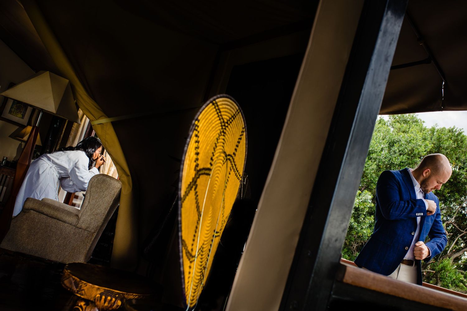Groom in blue suit adjusts tie on porch beside decorative yellow surfboard at outdoor wedding venue.