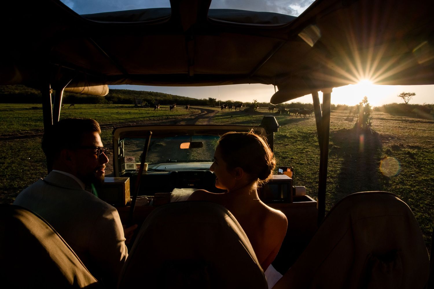 Couple riding in open safari vehicle at golden sunset, silhouetted against glowing sun rays over lush green landscape.