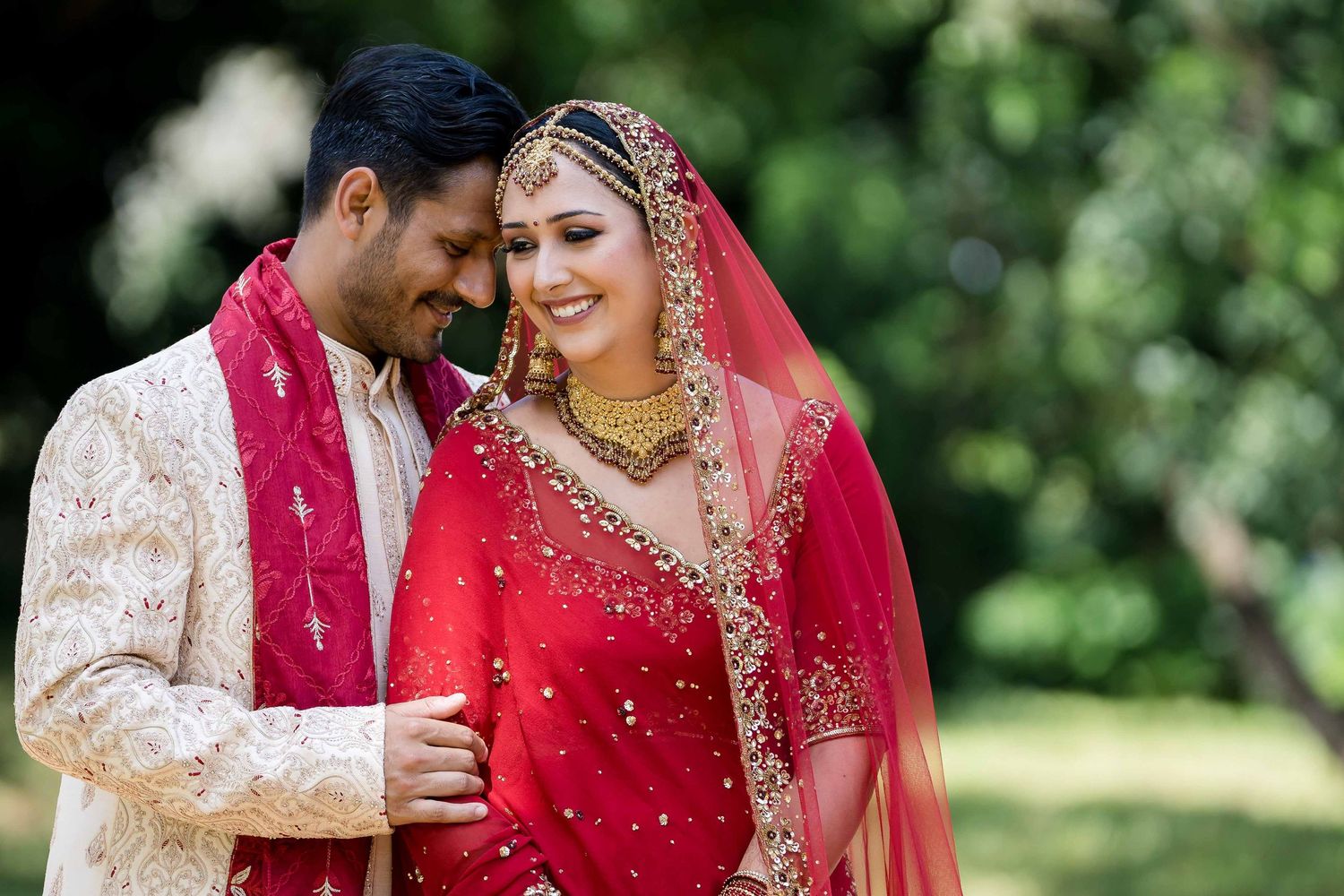 A couple in traditional Indian wedding attire shares joyful moments during an outdoor photo session in a garden setting.