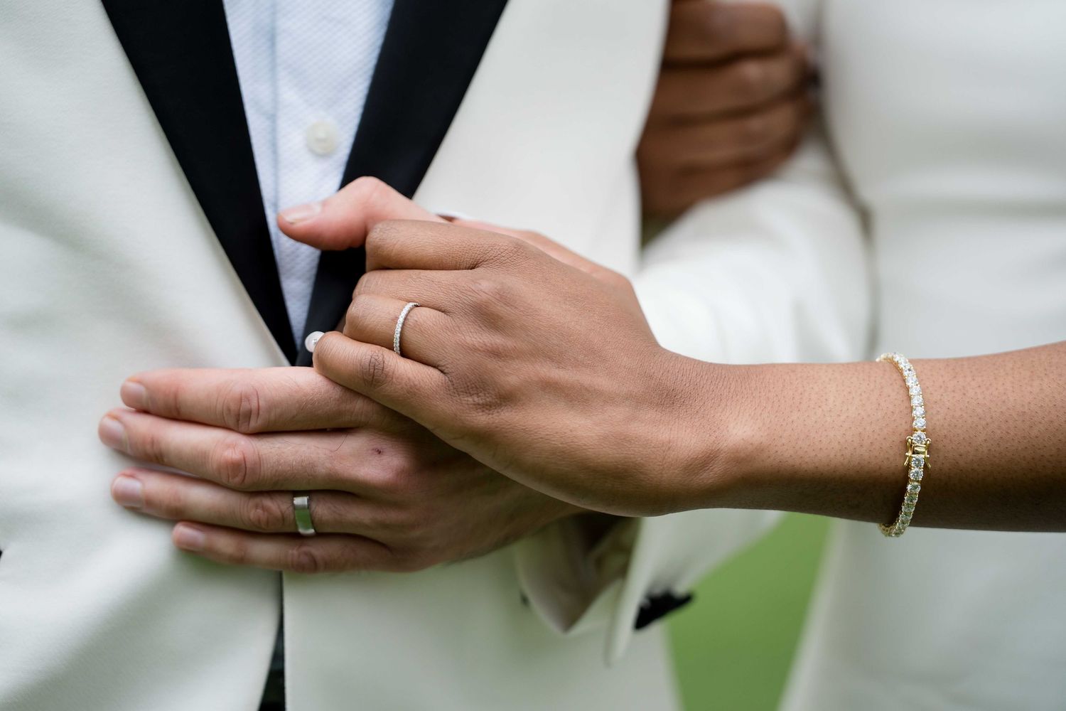 Close-up of hands wearing wedding rings and a delicate bracelet against black and white formal attire.