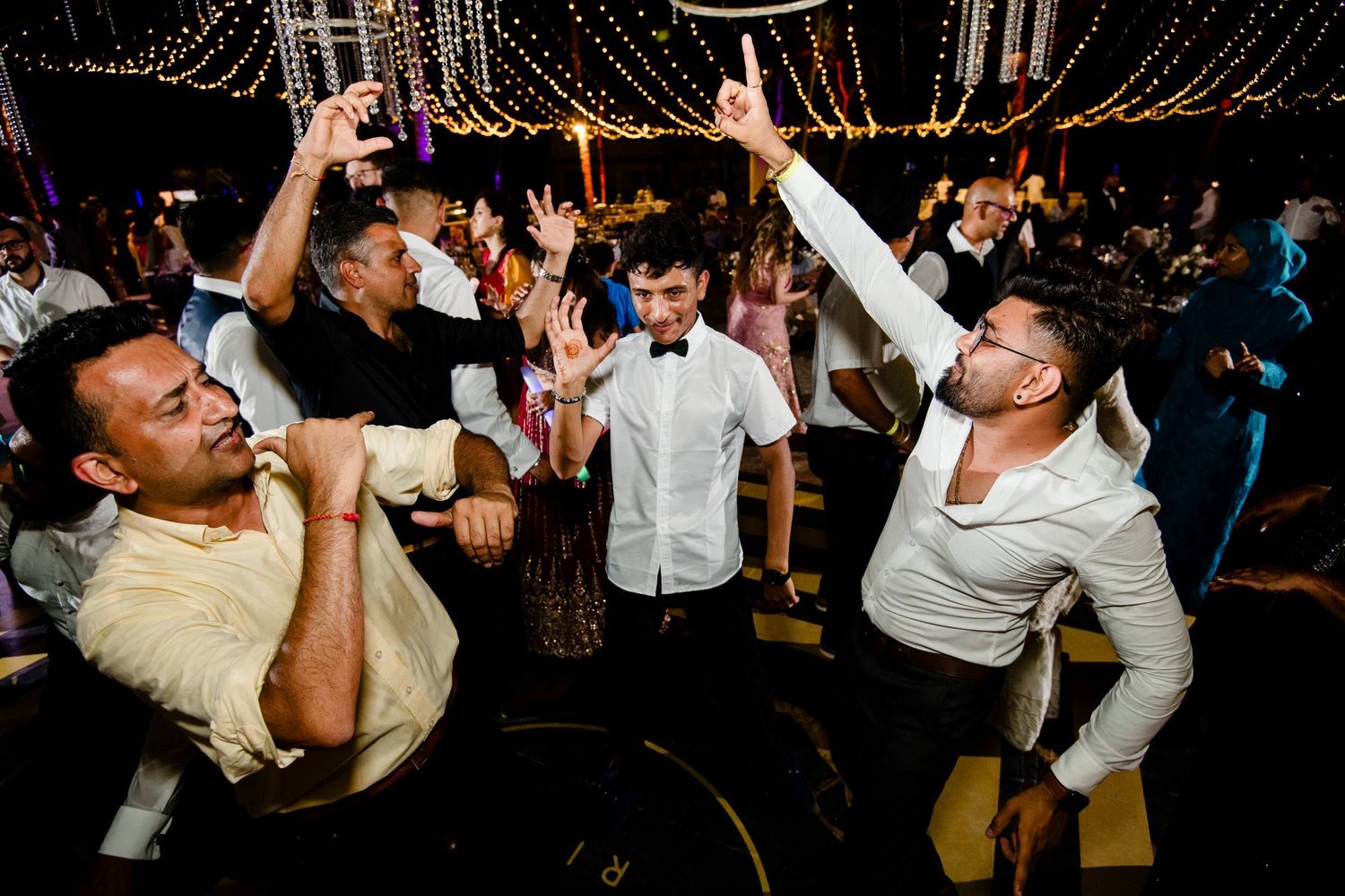 People dancing and celebrating at a lively nighttime wedding reception under decorative string lights.