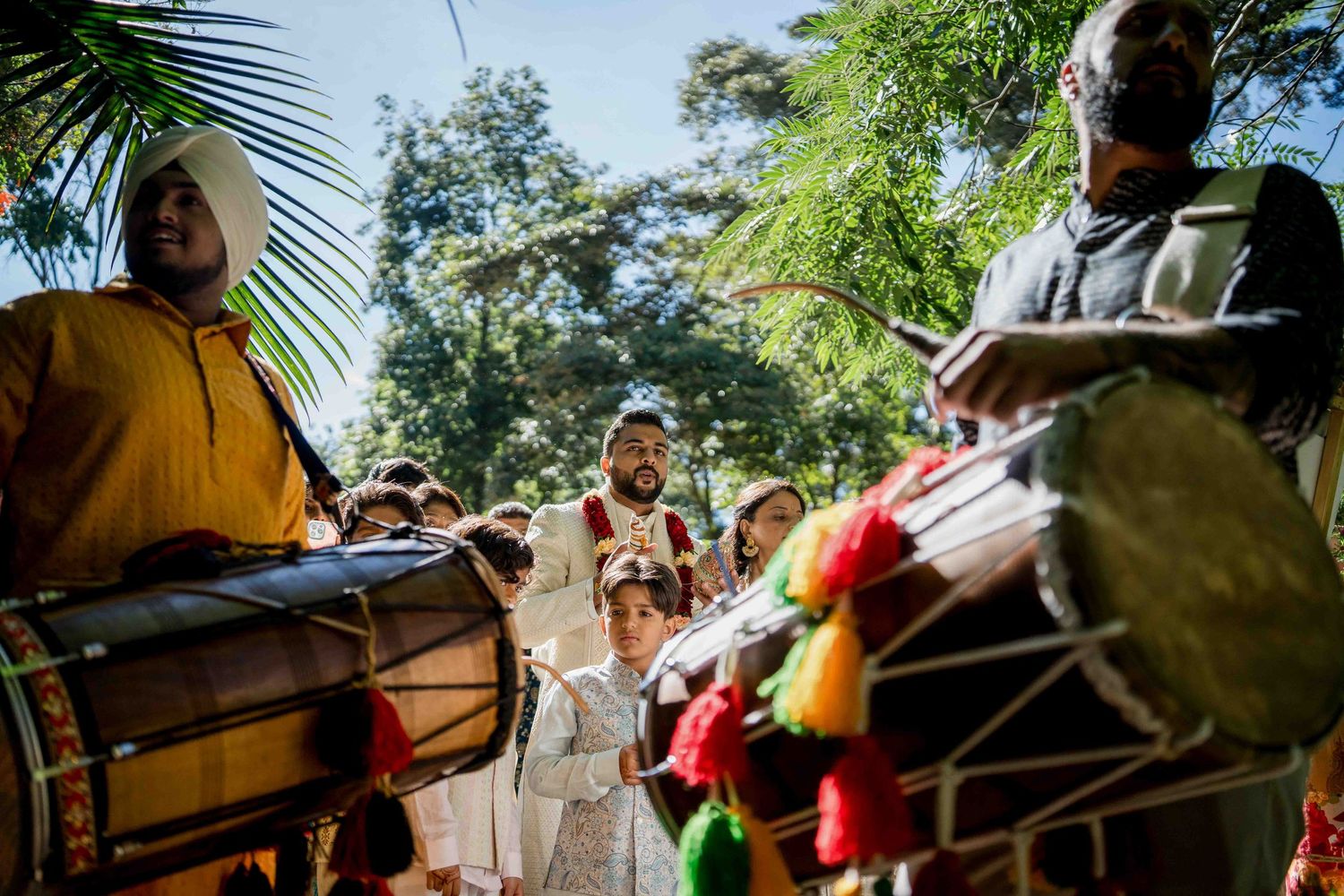 Traditional drums and musicians perform at an outdoor celebration with palm fronds overhead.