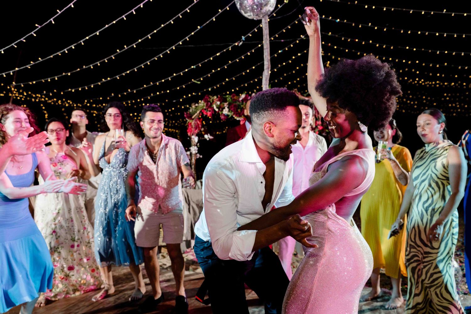 Wedding guests dance and celebrate under twinkling string lights at an outdoor evening reception.