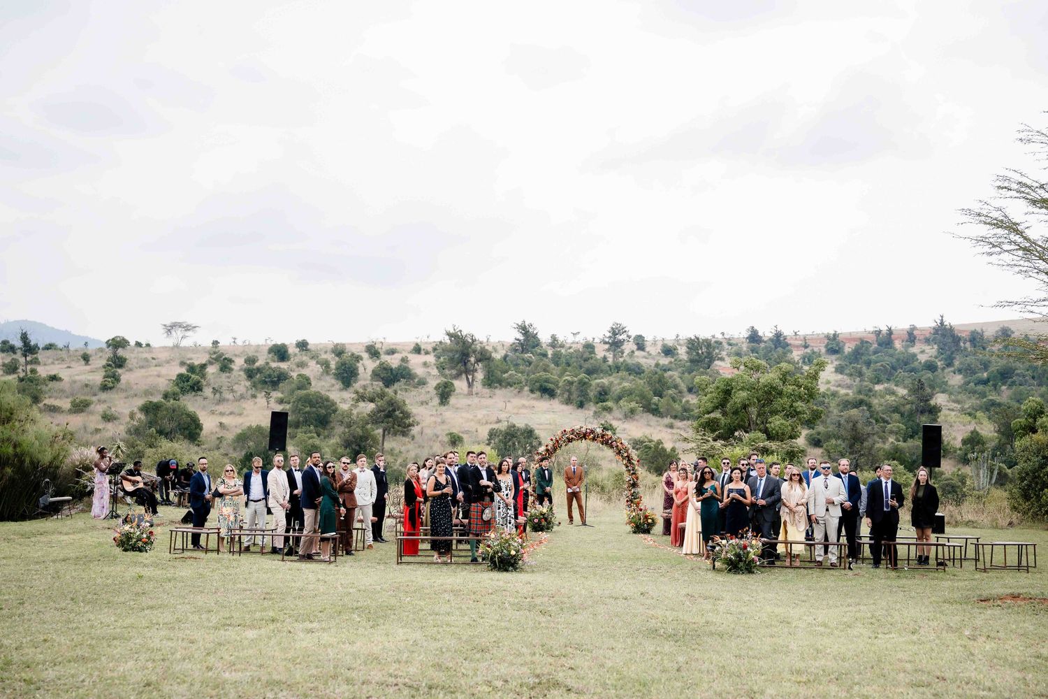 Wedding guests gather on a lush green hillside for an outdoor ceremony under a floral arch in a scenic natural setting.