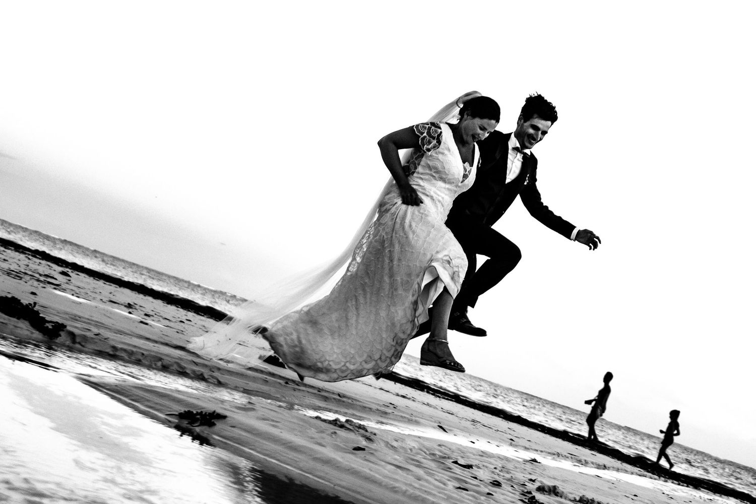 Dynamic black and white photo of couple running playfully on beach with children in background.