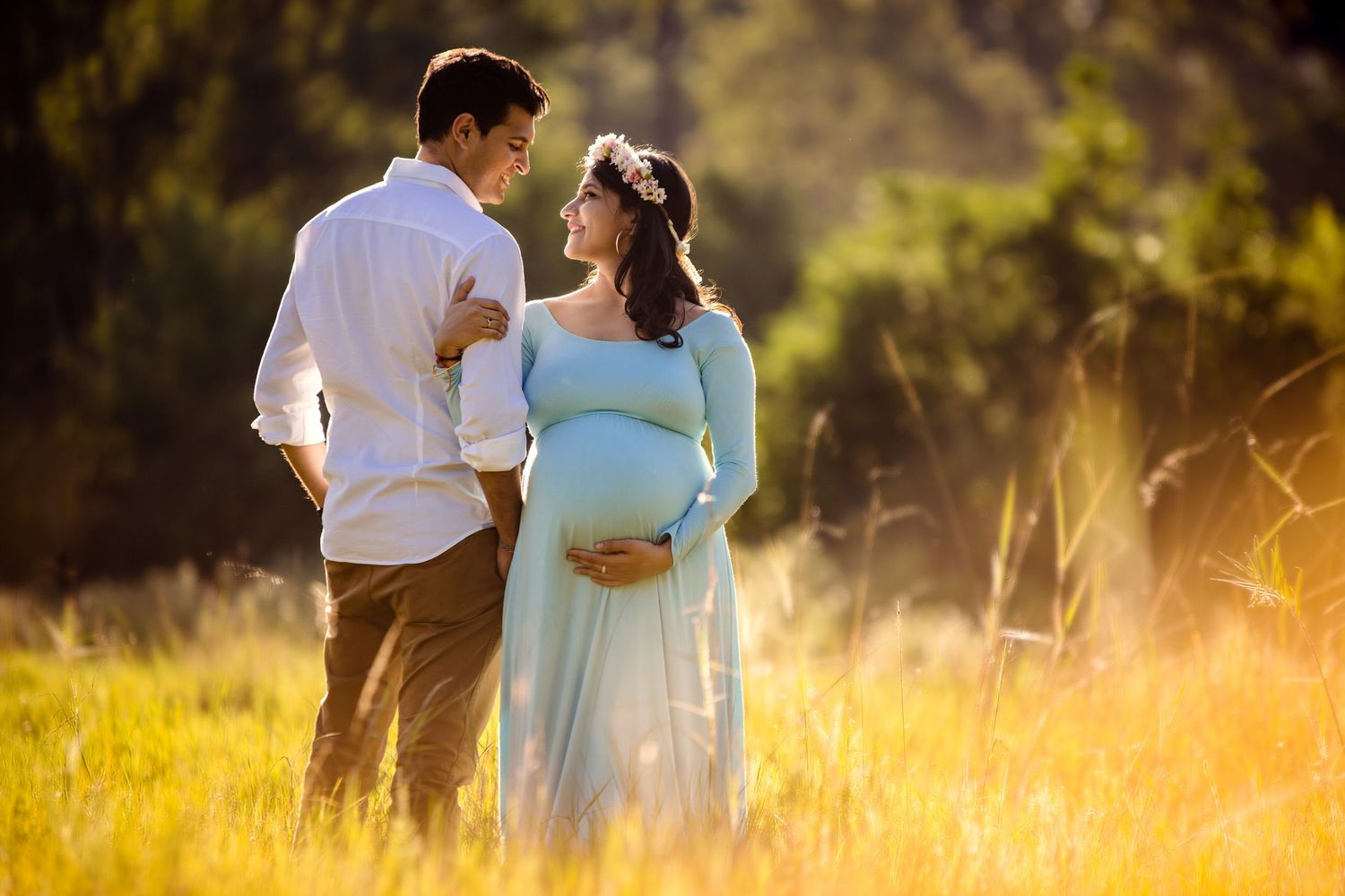Couple embraces in golden sunlit field wearing white and light blue clothing during romantic outdoor photoshoot.