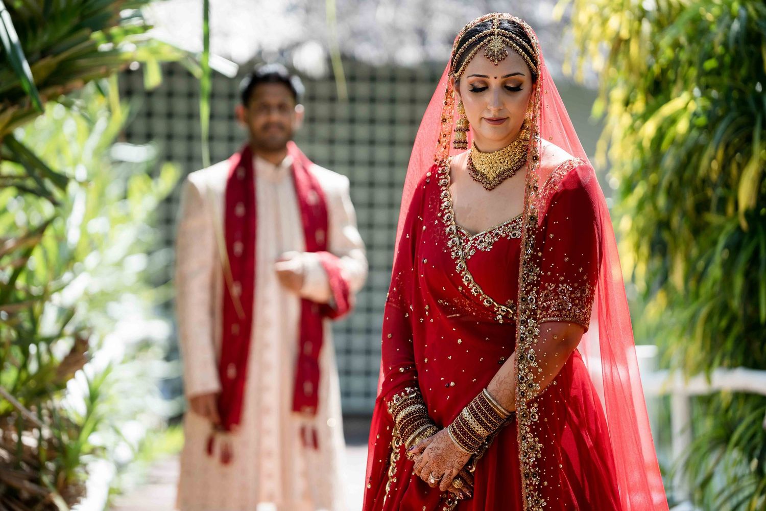 Traditional South Asian wedding couple in red and gold attire pose in a lush garden setting.