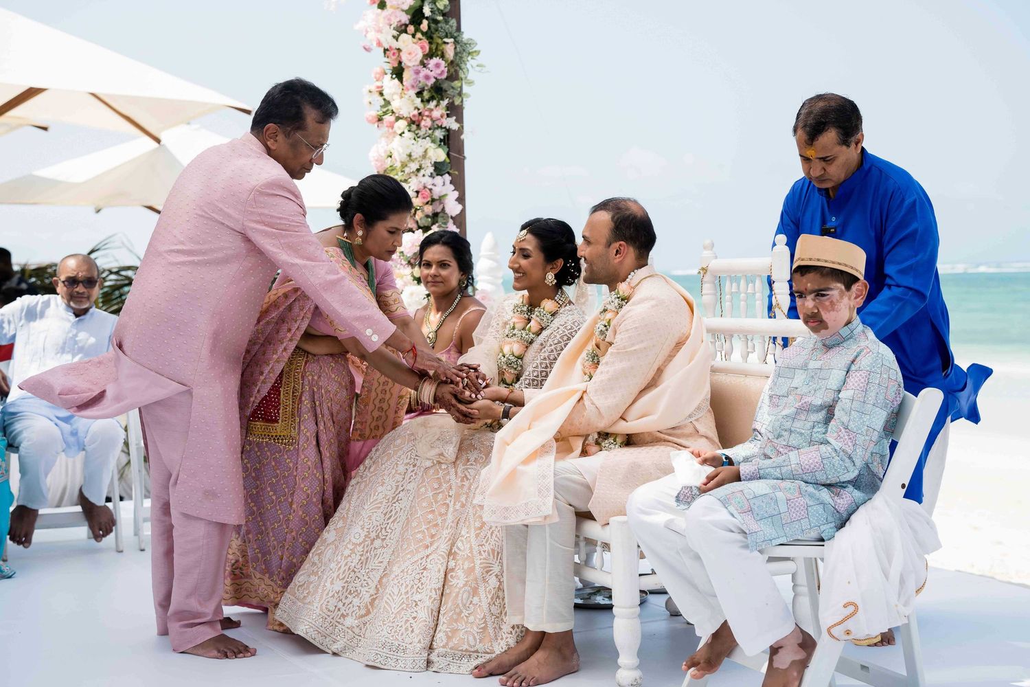 A traditional Indian wedding ceremony takes place on a white platform by the ocean with guests in pastel formal attire.
