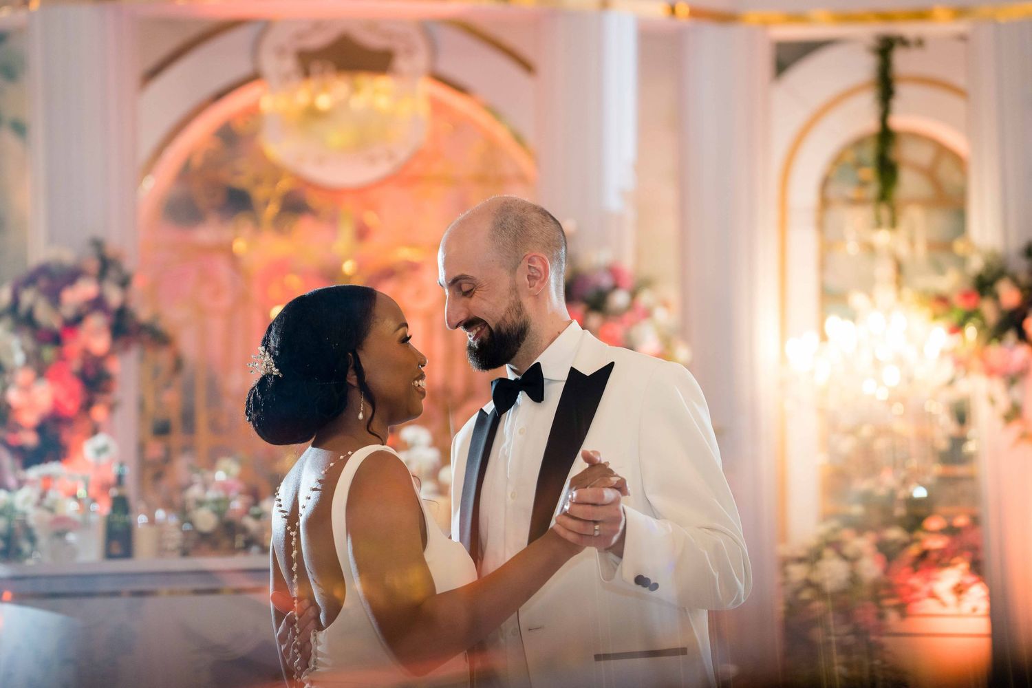 A wedding couple dances together in an elegant ballroom with warm lighting and floral decorations.