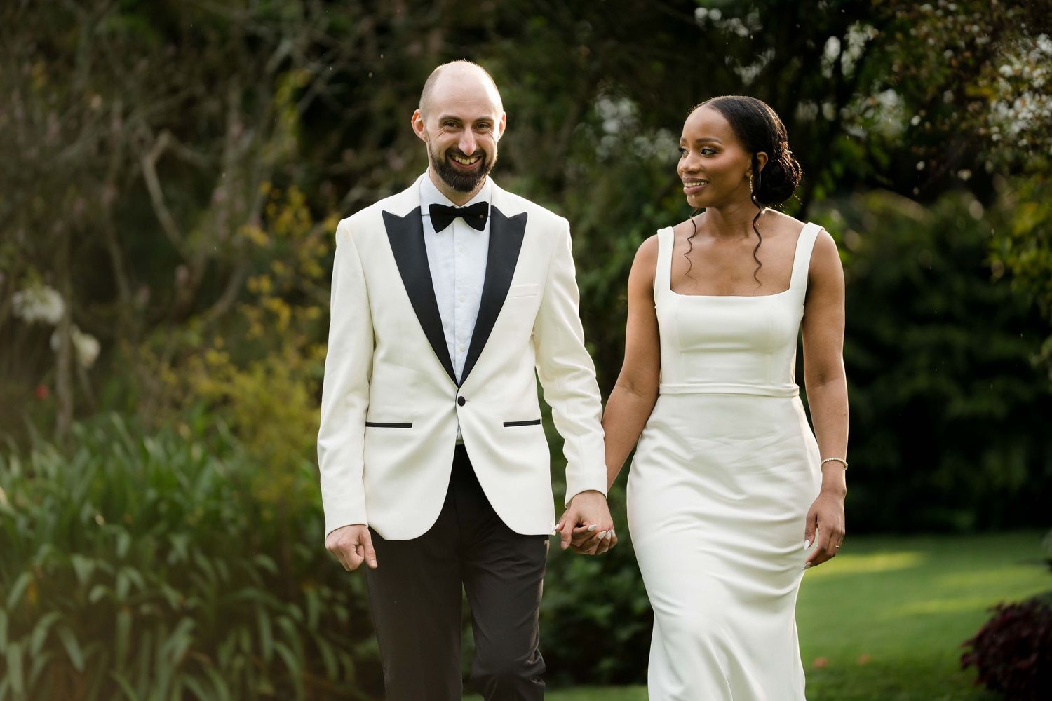 Couple in elegant wedding attire walking together and smiling on a lush green lawn.