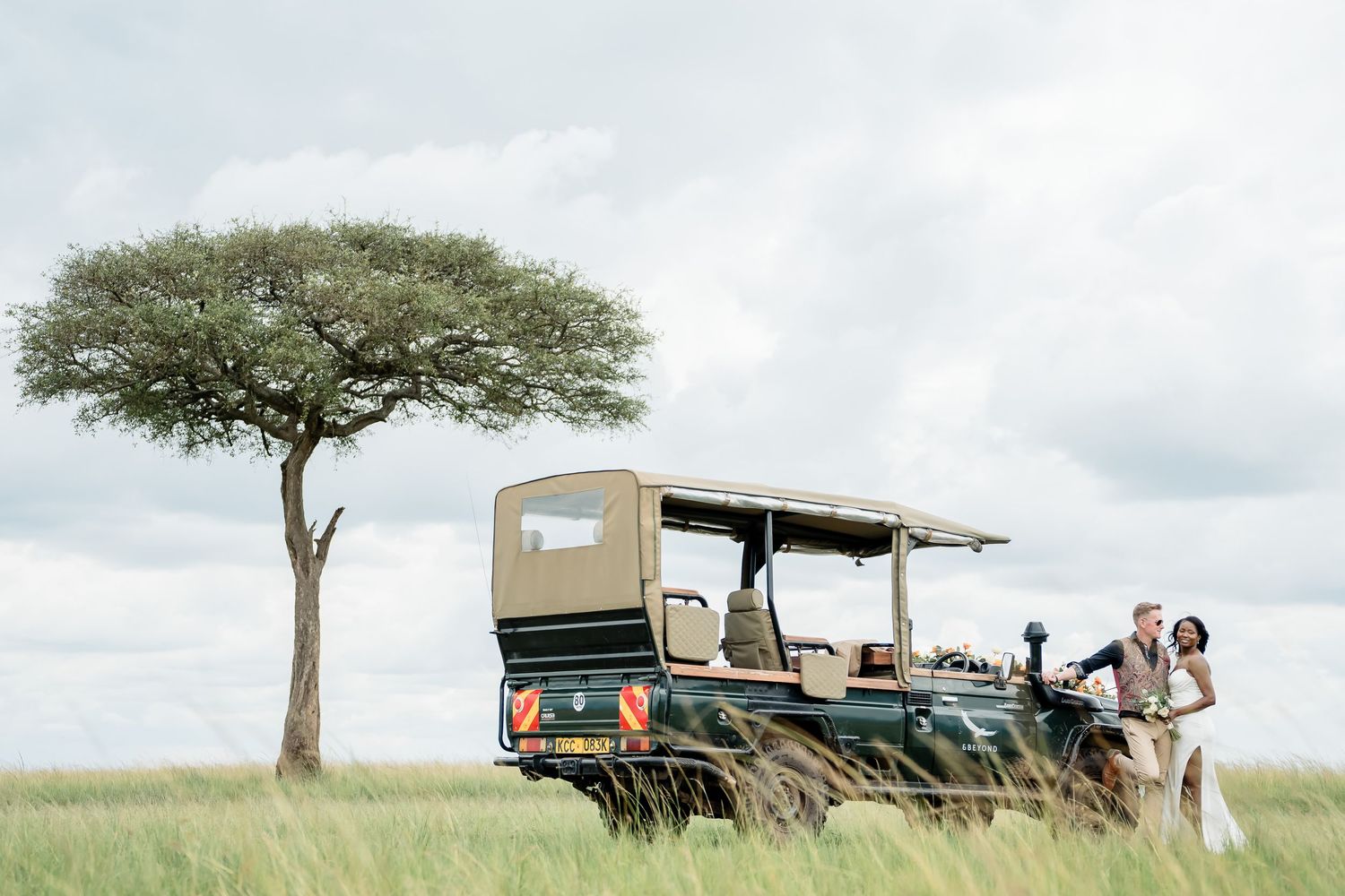 A safari vehicle parked beside an acacia tree in a vast African savanna during an overcast day.