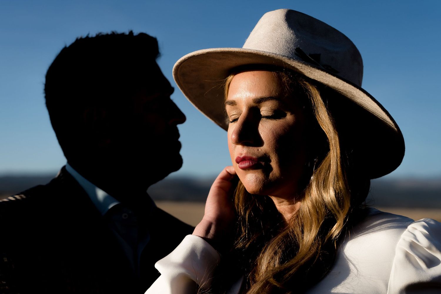 A woman in a wide-brim hat closes her eyes as a man in a suit leans close, set against a bright blue sky.