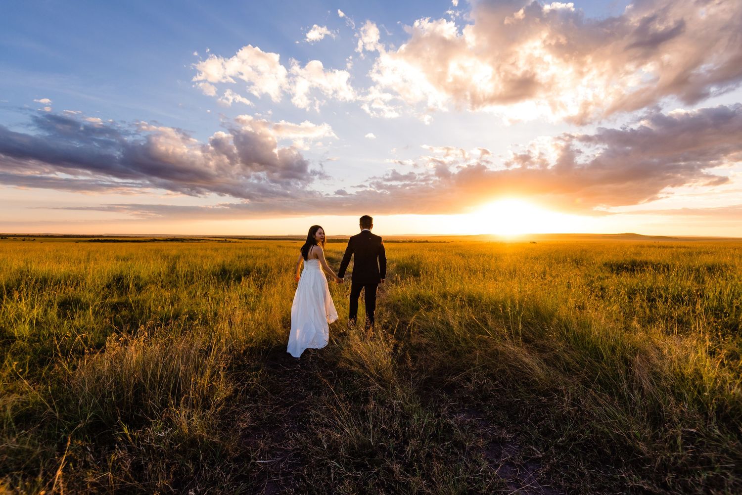 A couple walks through a golden wheat field at sunset with dramatic clouds overhead.