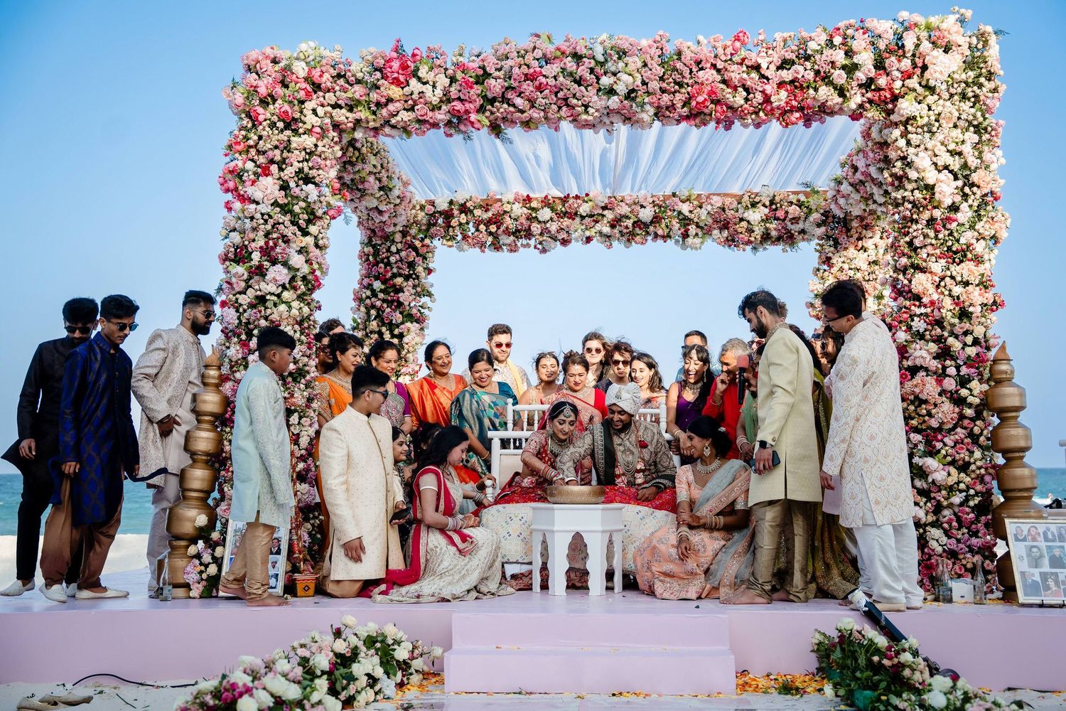 A wedding ceremony setup with a floral arch and guests gathered under a pink-tinted sky at an outdoor venue.