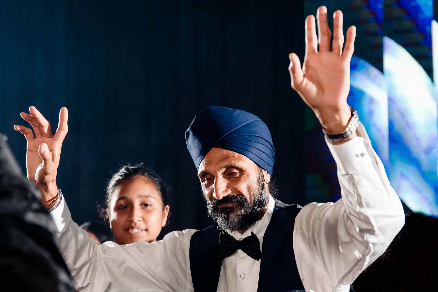 Wedding guests raise their hands in celebration during a festive reception with colorful lighting.