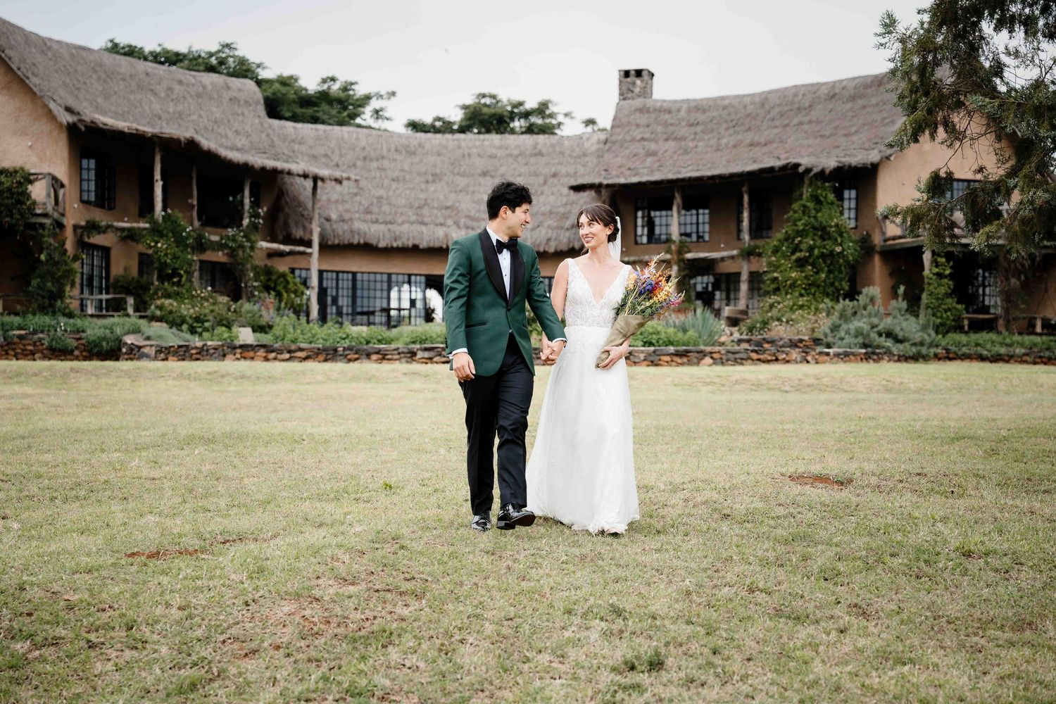 A couple in wedding attire walks together on a grassy lawn near rustic buildings on their special day.