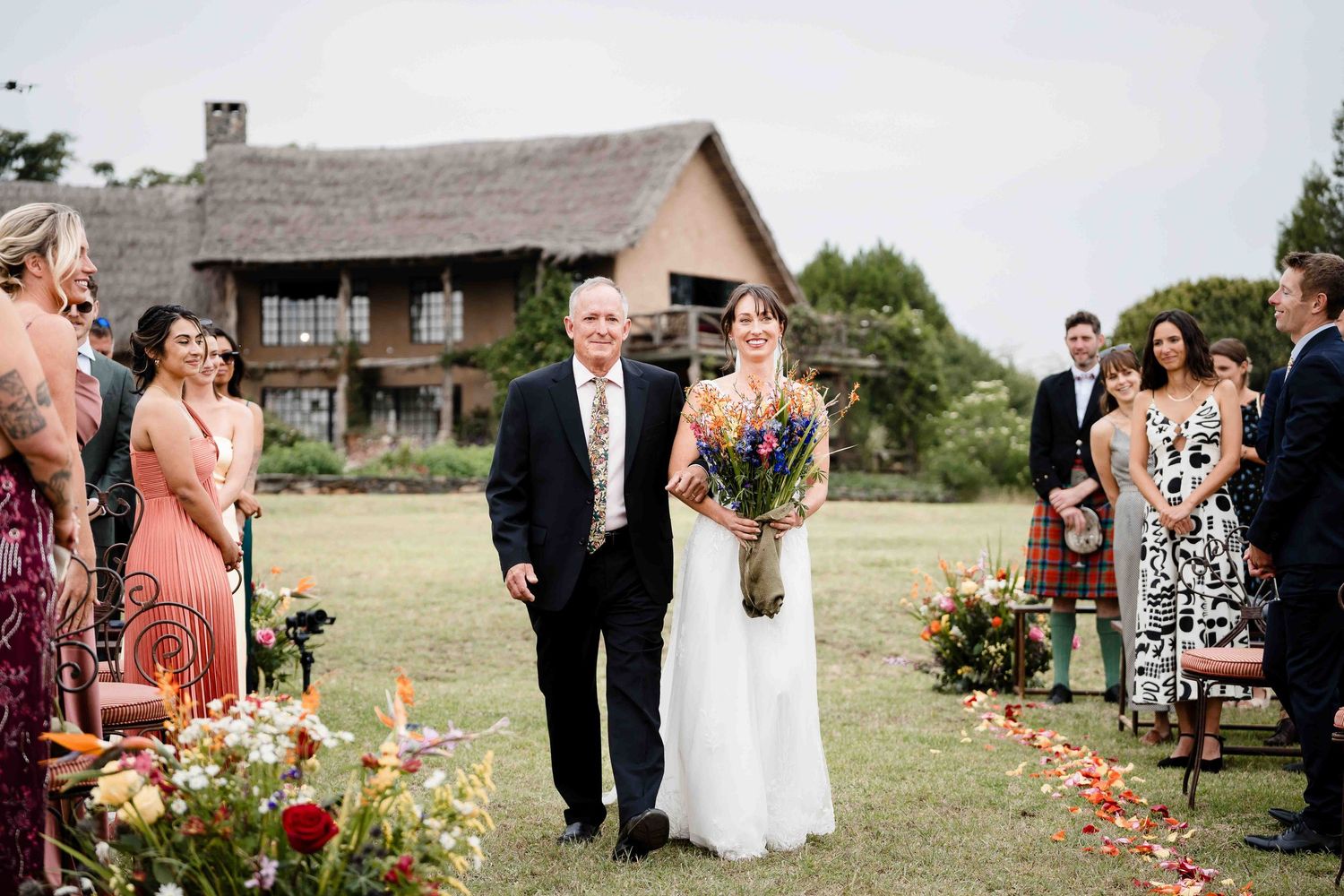 A rustic barn wedding ceremony with guests gathered outdoors in front of a wooden building on a grassy field.