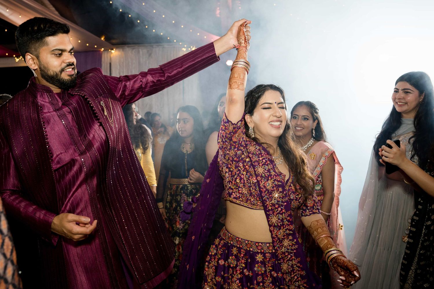 Dancers in burgundy traditional Indian attire celebrate during a festive event with sparkling lights.