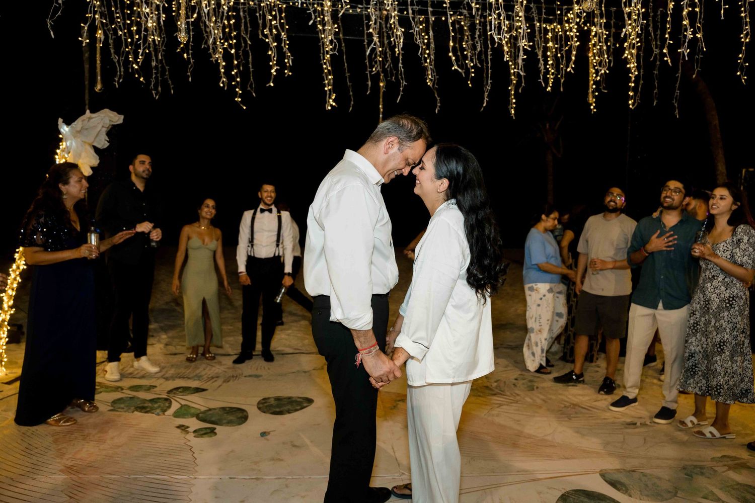 A couple dances together during a nighttime celebration under hanging string lights at an outdoor venue.