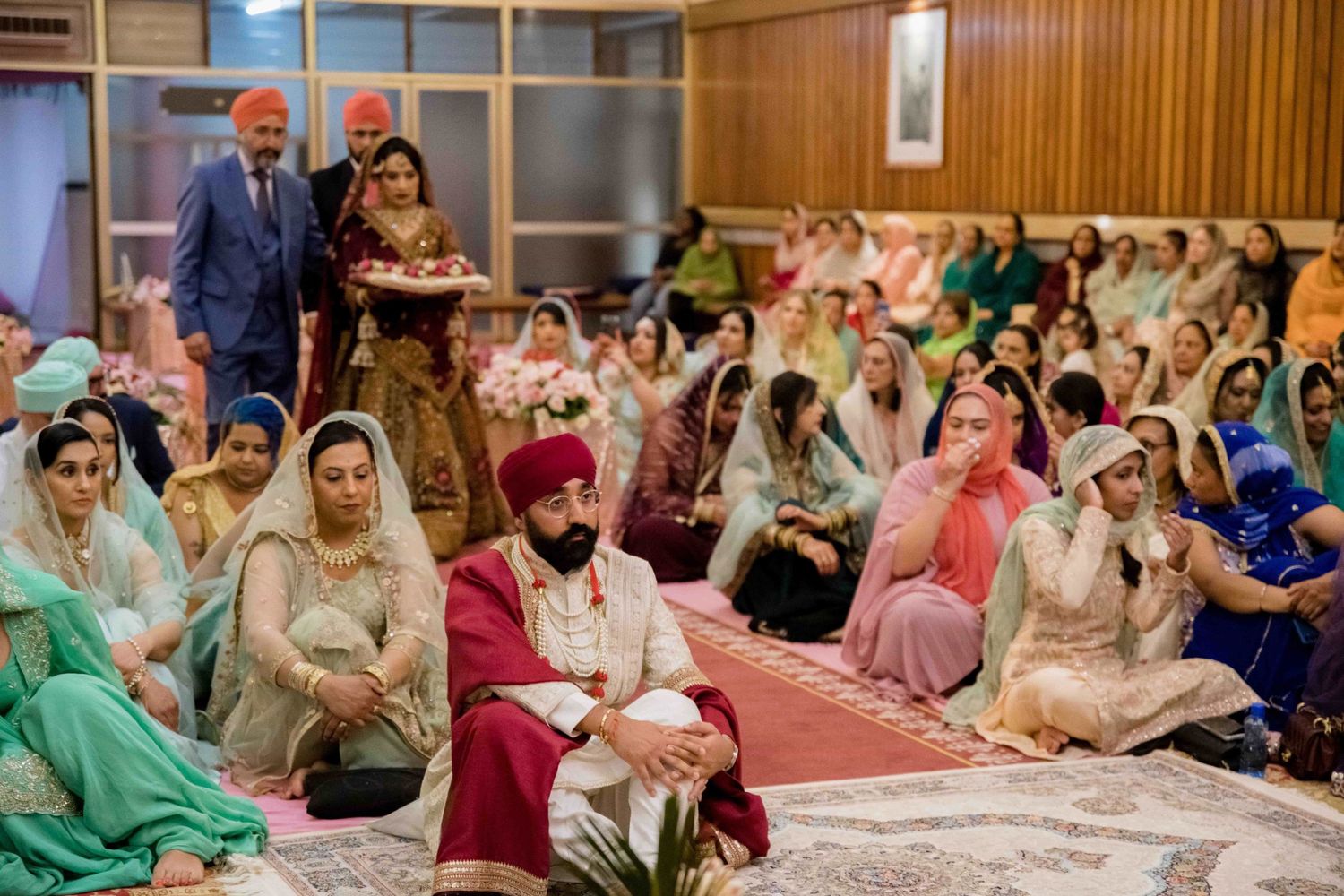 Guests seated on floor during traditional Sikh wedding ceremony in ornate indoor setting.