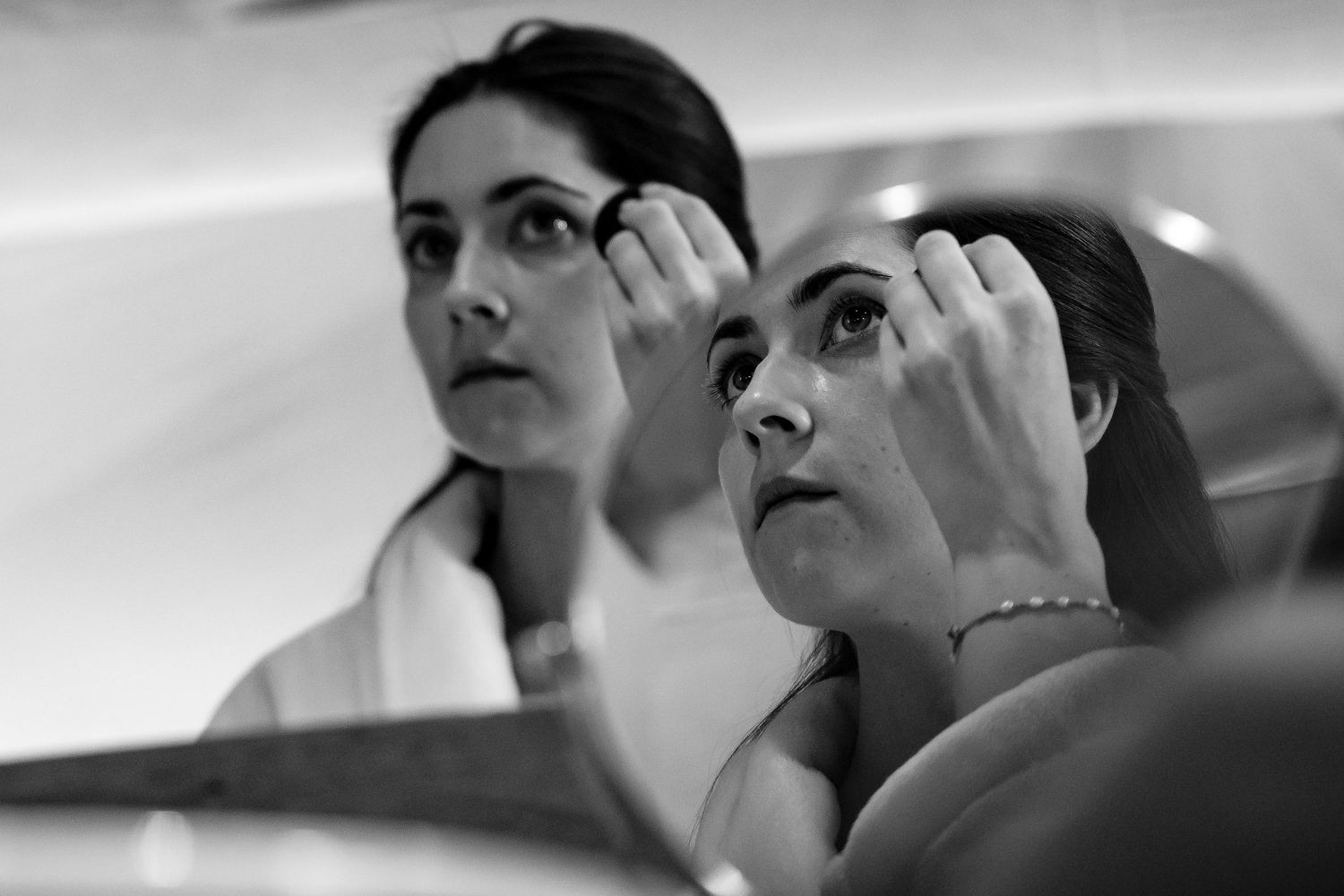 Two figures carefully apply makeup while looking in a mirror.