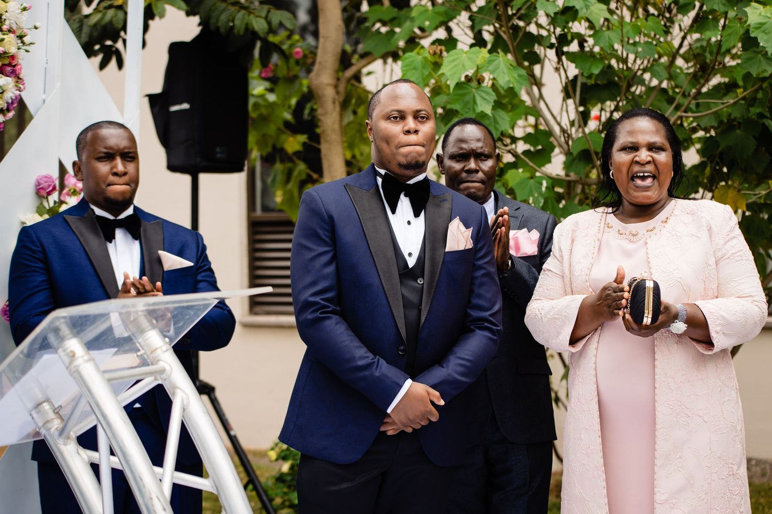 Wedding party in formal attire poses outdoors among garden greenery.
