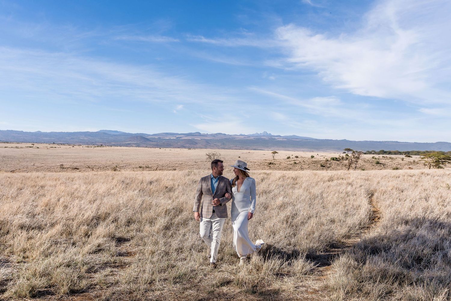 Couple walking through golden grasslands with mountain views and blue sky backdrop.