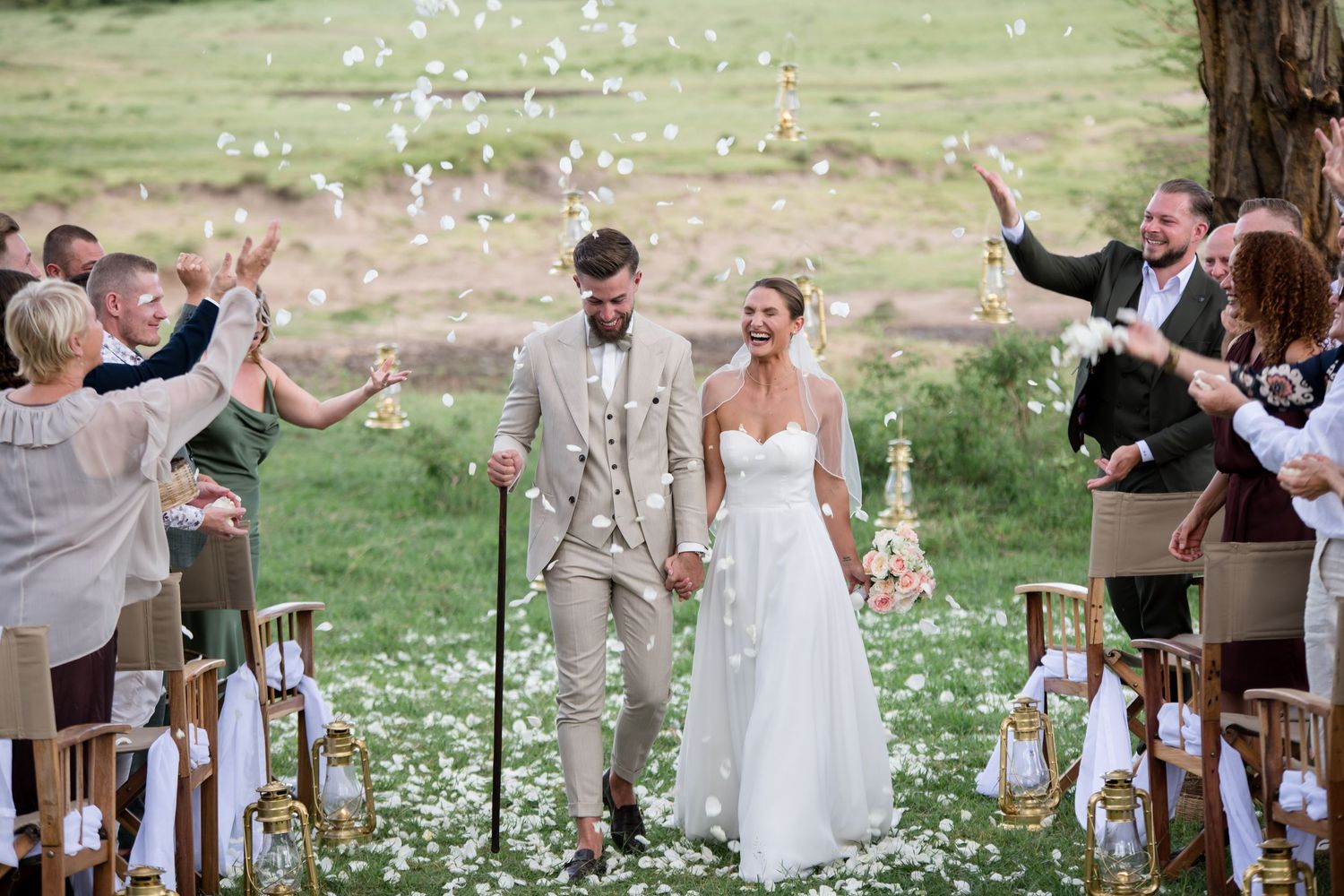 Couple walks down outdoor wedding aisle holding hands while guests toss white flower petals in celebration.