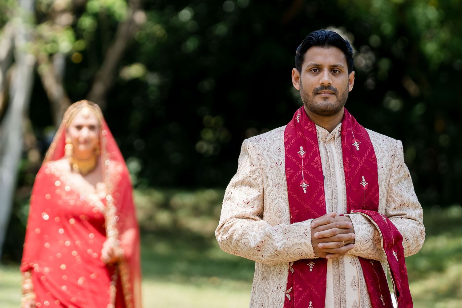 Traditional Indian wedding attire with cream sherwani and red dupatta in outdoor garden setting.