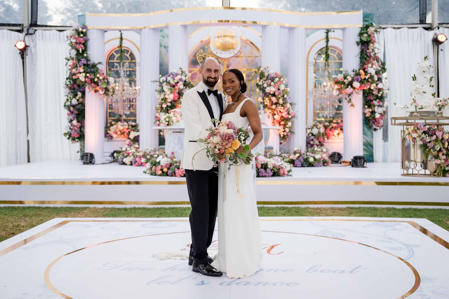 A couple dances together in an elegant white venue decorated with floral arrangements and romantic lighting.