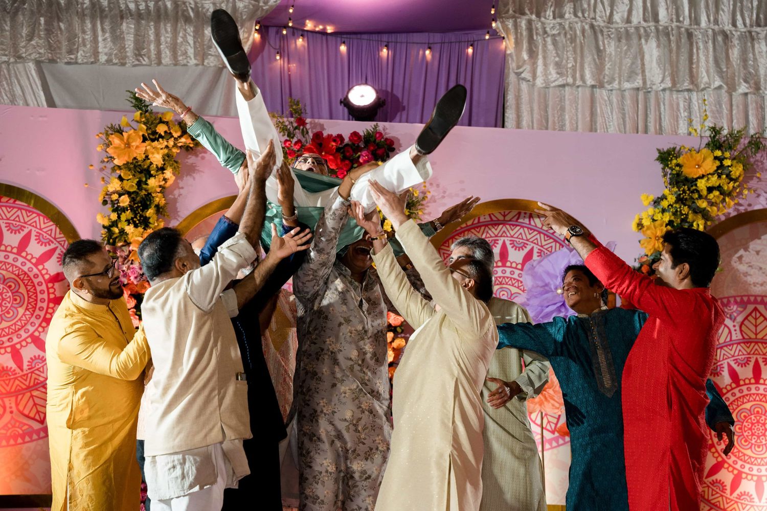 Celebrants lift their hands in joy during a festive Indian wedding celebration with decorative backdrop.