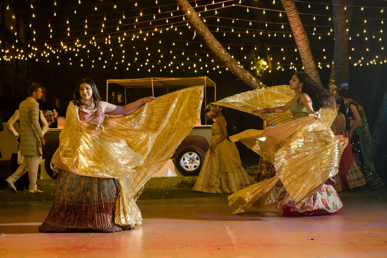 Dancers in flowing gold dresses perform under twinkling lights at an outdoor event.