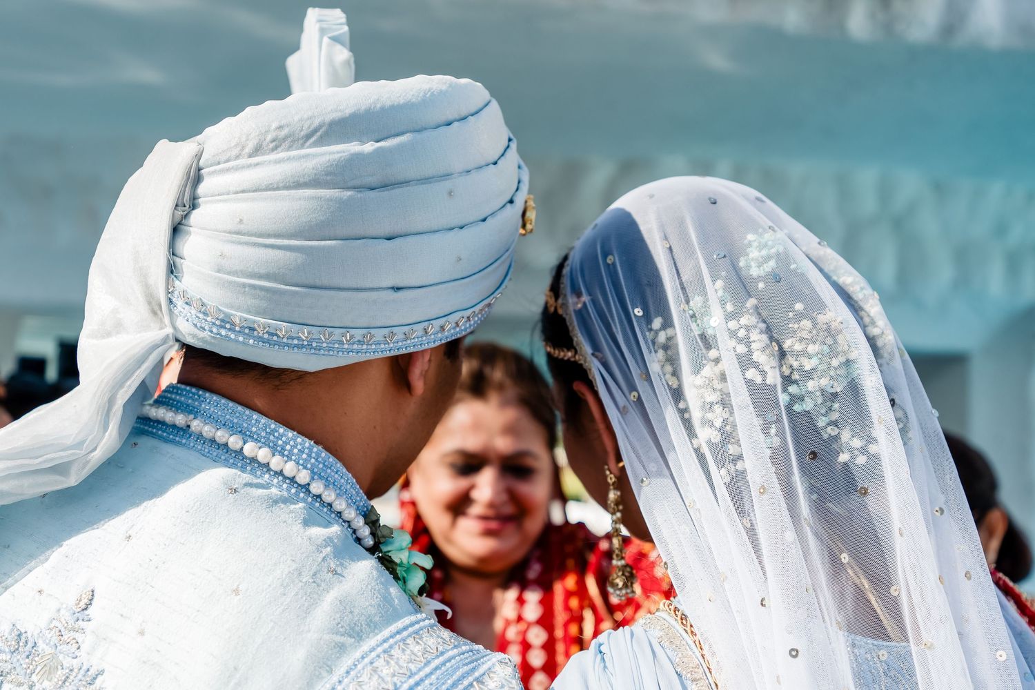 Couple at a traditional Indian wedding ceremony with white turban and veil against a turquoise background.